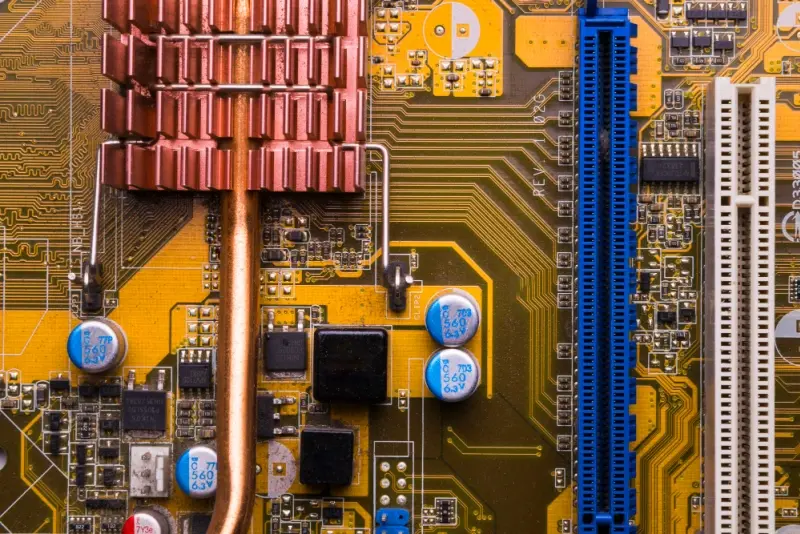 Substrate Material Selection: close-up of a motherboard showing a copper heat pipe with finned heatsink, blue expansion slots, radial capacitors, and dense yellow PCB traces.